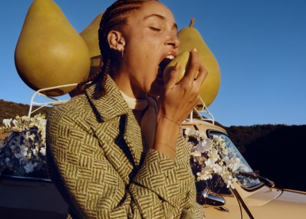 Adwoa Aboah con pelo trenzado, mordiendo una pera con un fondo de tres grandes peras de atrezo encima del portaequipajes de un coche.