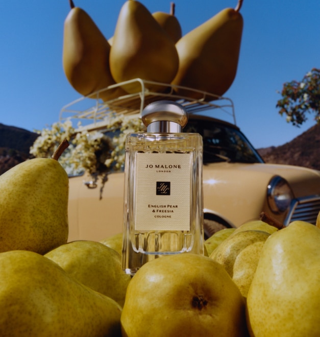 Frasco de Cologne English Pear & Freesia de 100 ml rodeado de peras frescas con un fondo de tres grandes peras de atrezo encima del portaequipajes de un coche.